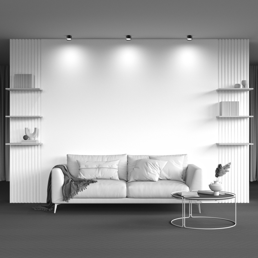 Svetlá pohovka stojí pred bielou stenou v obývacej izbe. Na pohovke sú vankúše a deka. Vedľa pohovky je nízky odkladací stolík. Po stranách steny sú úzke police s knihami a dekoráciami.