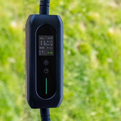 Nabíjacia stanica pre elektromobily zobrazuje údaje: 15,5 A, 244 V, 3,1 kW, 0,1 kWh, 00:00. Zariadenie je čierne, visí vonku na zelenom pozadí.
