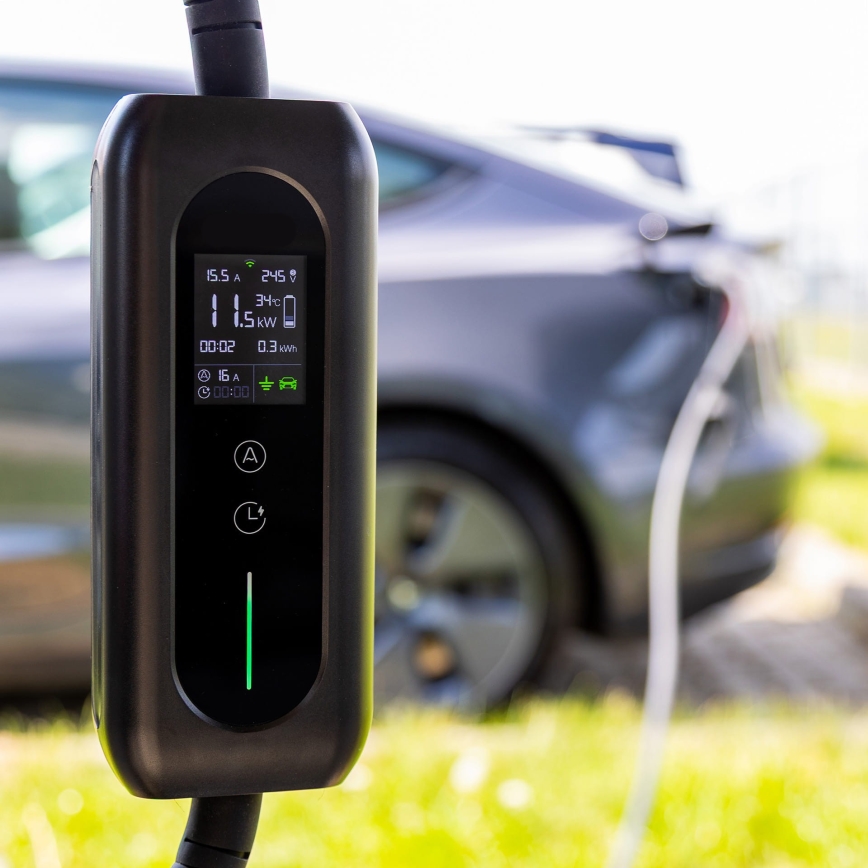 Nabíjacia stanica pre elektromobily nabíja auto. Na displeji sa zobrazuje: 15,5 A, 245 V, 1,5 kW, 3°C, 00:02, 0,3 kWh, 15 A, 00:00. Kontext: vonku, trávnatá plocha.
