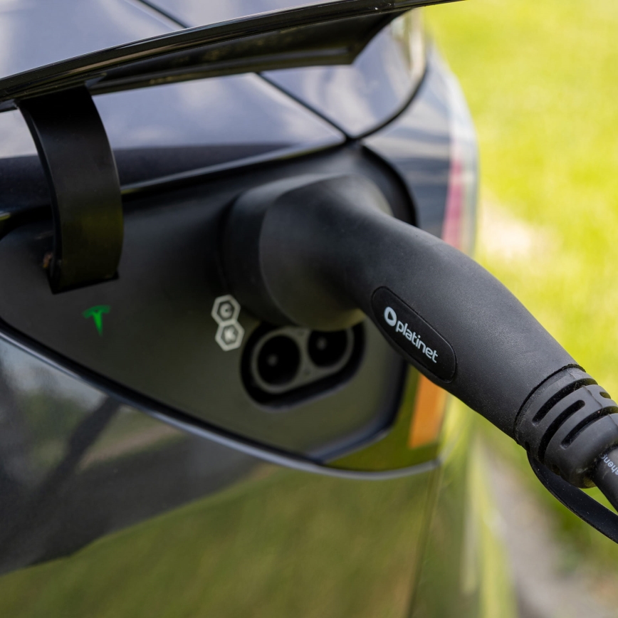 Elektrické auto sa nabíja. Nabíjací kábel s označením „oplanet“ je zasunutý do nabíjacej zásuvky auta. Auto stojí vonku na tráve.