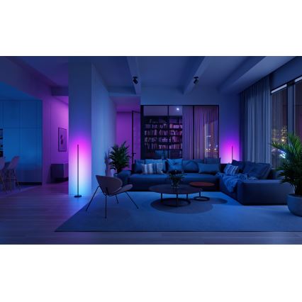 Moderná podlahová lampa s farebným LED osvetlením svieti v obývacej izbe s pohovkou, konferenčným stolíkom a rastlinami. Atmosféra je tlmená, prevažujú modré a fialové tóny.