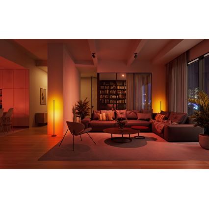 Stĺpová lampa svieti v oranžovom svetle v modernej obývacej izbe s hnedou sedačkou, konferenčným stolíkom a rastlinami. Izba je osvetlená tlmeným, teplým svetlom.