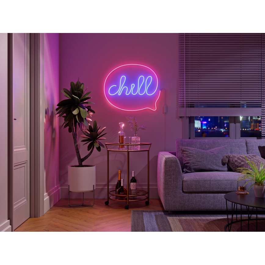 Neonové svetlo v tvare bubliny s nápisom „chill“ svieti na stene. Je umiestnené v obývacej izbe s pohovkou, stolíkom a izbovými rastlinami.