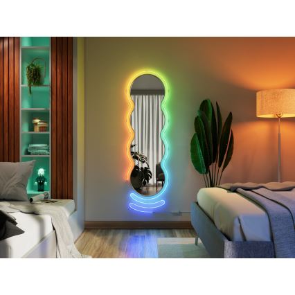 Zrkadlo s vlnitým okrajom osvetlené dúhovými LED diódami visí na stene spálne. Zrkadlo odráža časť miestnosti. Okolím sú posteľ, lampa a rastlina.