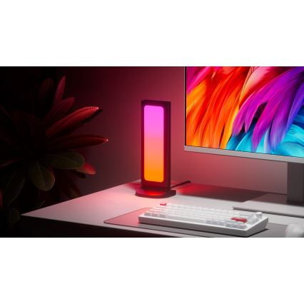 Svietiaca lampa s prechodom farieb od ružovej po oranžovú stojí na stole popri počítači s klávesnicou. Lampa je umiestnená na tmavom podstavci. V pozadí je rastlina a monitor s farebnou grafikou.