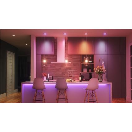 Kuchynská linka s tromi stoličkami je osvetlená farebnými LED svetlami, stropnými svietidlami a LED pásom pod linkou. Svetlá majú rôzne odtiene ružovej a oranžovej. V pozadí sú kuchynské skrinky a drevená stena.