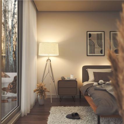 Stojacia lampa s textilným tienidlom svieti v spálni. Vedľa nej je nočný stolík s knihou a hrnčekom. V pozadí je posteľ s posteľnou bielizňou a oknom s výhľadom na vonkajšiu scénu.