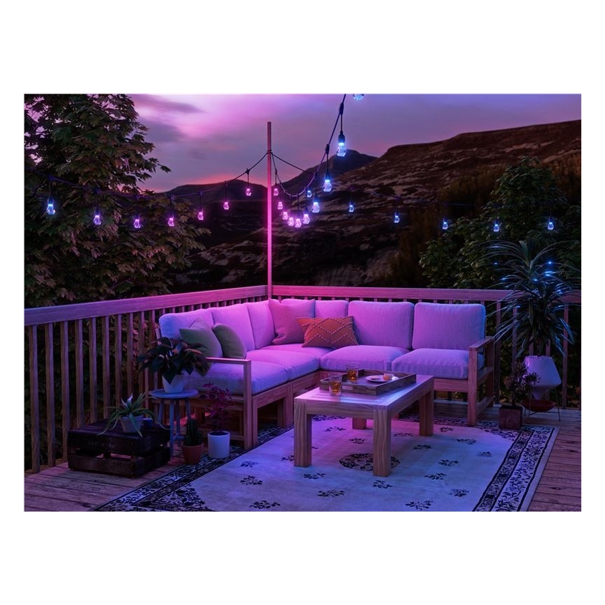 Rohová sedačka s nízkym stolíkom sa nachádza na drevenej terase s farebným osvetlením. Okolí zdobia rastliny a girlanda s LED žiarovkami svietiacimi v ružovej a modrej farbe. V pozadí je vidieť kopce a zapadajúce slnko.