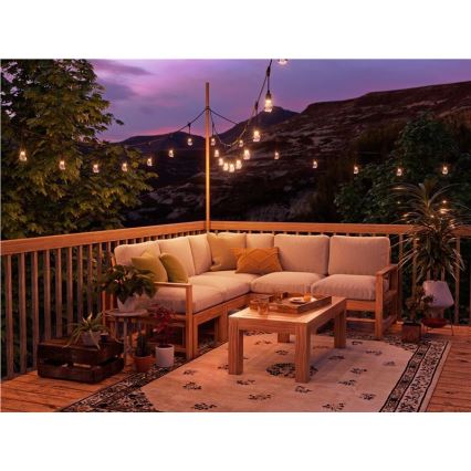 Rohová záhradná sedačka stojí na drevenom balkóne s kobercom. Na balkóne je aj nízky drevený stolík. Nad balkónom sú rozvešané svetelné reťaze. V pozadí sú viditeľné kopce pri západe slnka.