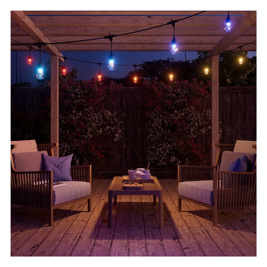 Farebné svetielka osvetľujú vonkajšiu terasu. Svietia z vonkajšieho osvetlenia zaveseného na drevenej pergole. Na terase sa nachádzajú dve kreslá a nízky stolík.