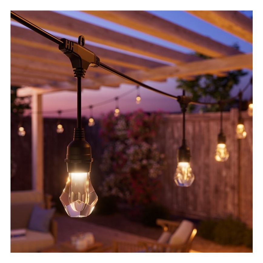 Svetlá svietia v exteriéri. Viacero ozdobných žiaroviek visí na vonkajšom elektrickom vedení pod drevenou pergolou. V pozadí je záhrada s nábytkom.