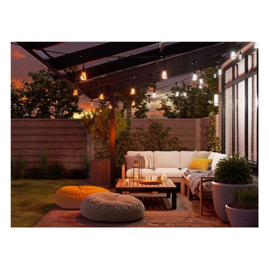 Svietidlá svietia na vonkajšej terase. Záhradný nábytok, vrátane pohovky a taburetky, je umiestnený na drevenom nábytku. V pozadí sa nachádza drevený plot a zeleň.