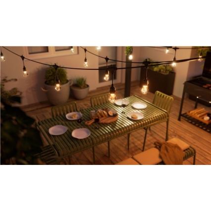 Vonkajšie osvetlenie s mnohými žiarovkami visí nad zeleným záhradným stolom s jedlom a stoličkami. Stojí na drevenej terase, vedľa je gril.