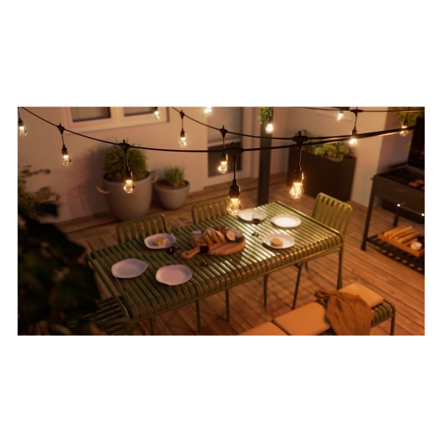 Vonkajšie osvetlenie s mnohými žiarovkami visí nad zeleným záhradným stolom s jedlom a stoličkami. Stojí na drevenej terase, vedľa je gril.