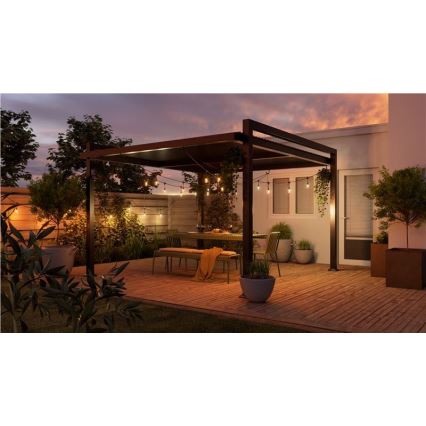 Záhradná pergola s osvetlením je umiestnená na drevenej terase. Stojí pod ňou záhradný nábytok – stôl a lavice. Okolie tvorí zeleň a časť domu.