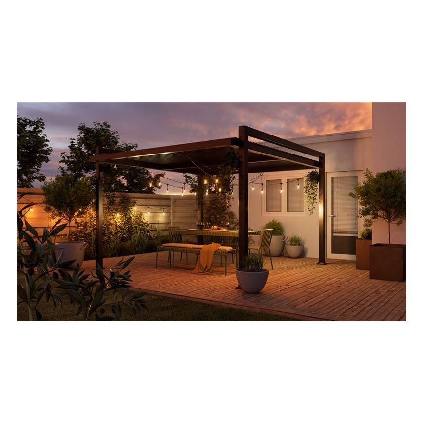 Záhradná pergola s osvetlením je umiestnená na drevenej terase. Stojí pod ňou záhradný nábytok – stôl a lavice. Okolie tvorí zeleň a časť domu.