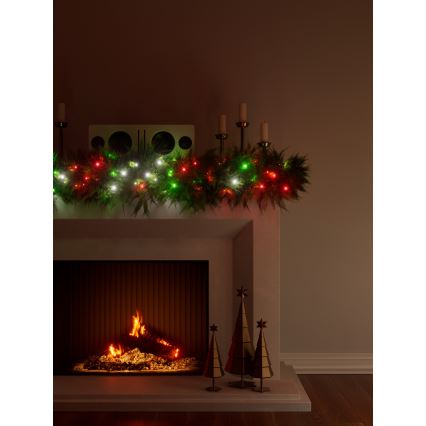 Vianočná girlanda so zelenými, červenými a bielymi svetielkami je umiestnená na kachľovej rímske. Pod ňou je krb s horiacim ohňom a tri zlaté ozdobné stromčeky.