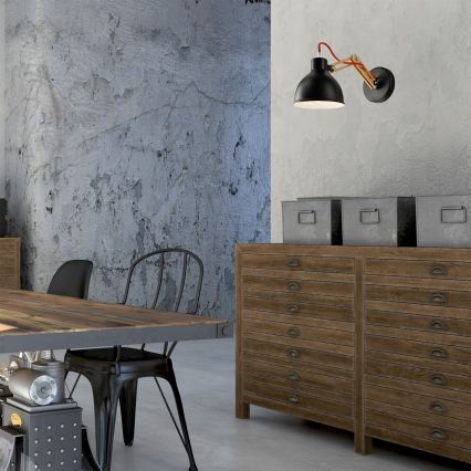 Čierna nástenná lampa s dreveným ramenom a červenou šnúrou svieti v industriálne zariadenej miestnosti. Lampa je umiestnená na stene s betónovou textúrou. V miestnosti sa nachádza drevený stôl a komoda s mnohými zásuvkami.