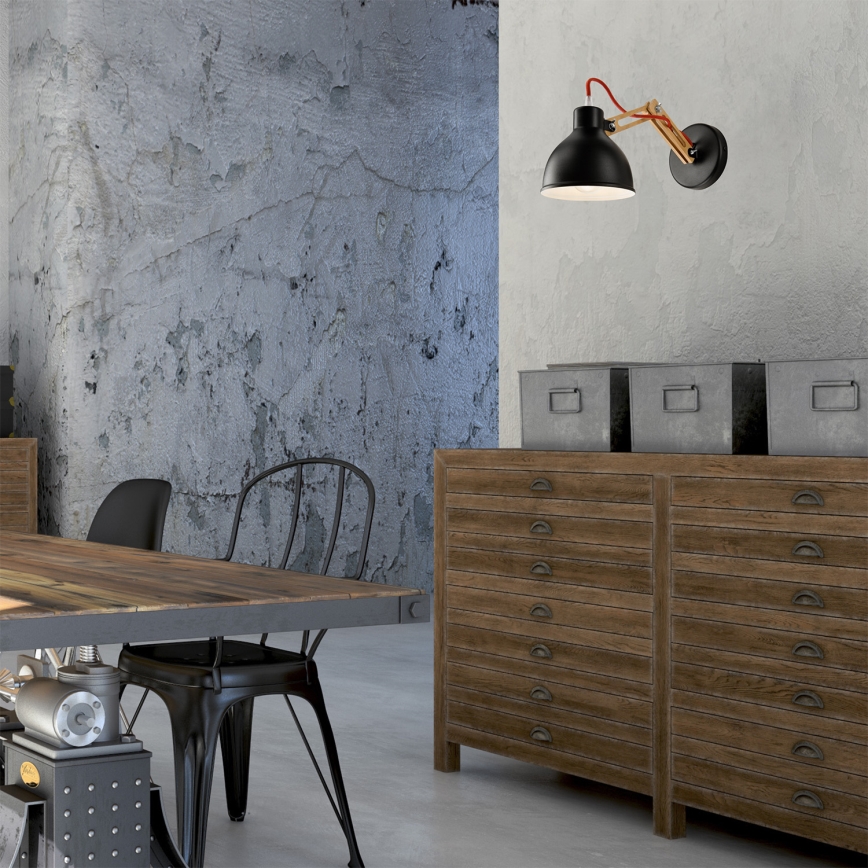 Čierna nástenná lampa s dreveným ramenom a červenou šnúrou svieti v industriálne zariadenej miestnosti. Lampa je umiestnená na stene s betónovou textúrou. V miestnosti sa nachádza drevený stôl a komoda s mnohými zásuvkami.