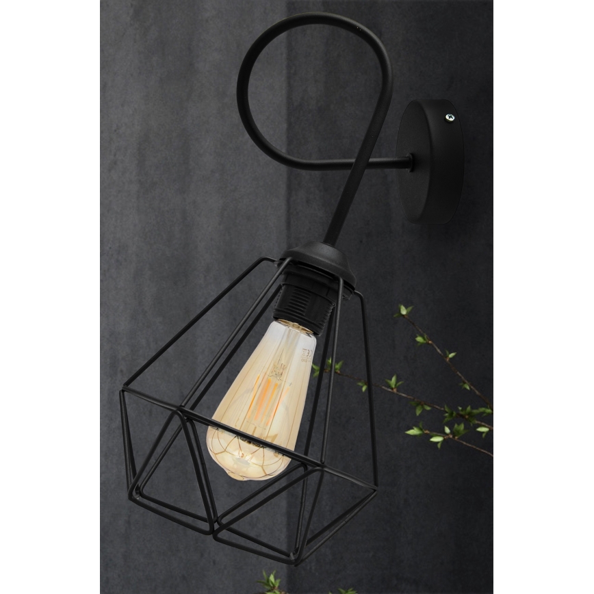Čierna kovová nástenná lampa s geometrickým tienidlom osvetľuje priestor. Lampa je pripevnená na stenu. V pozadí je tmavá stena a vetvička s listami.