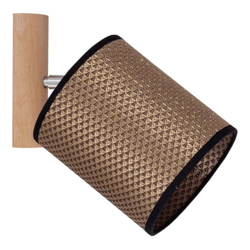 Nástenná lampa s valcovým tienidlom zo zlatej textílie s tmavou lemovkou, pripevnená k drevenej konštrukcii. Lampa je pripevnená na stene a smeruje dopredu.