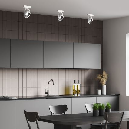 Tri stropné svietidlá svietia v moderne zariadenej kuchyni s tmavosivými skrinkami a svetloružovými dlaždicami. Stôl s čiernymi stoličkami sa nachádza pred kuchynskou linkou.