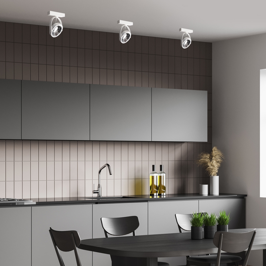 Tri stropné svietidlá svietia v moderne zariadenej kuchyni s tmavosivými skrinkami a svetloružovými dlaždicami. Stôl s čiernymi stoličkami sa nachádza pred kuchynskou linkou.