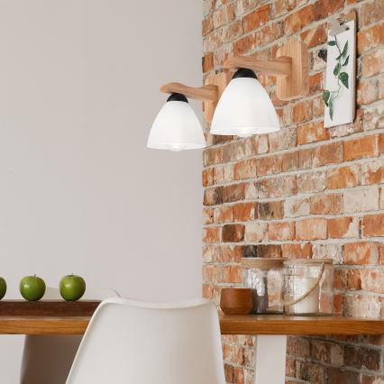 Drevená nástenná lampa s dvoma bielymi tienidlami je pripevnená k tehlovej stene. Lampa je umiestnená v jedálni pri stole.