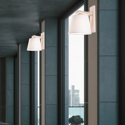 Nástenná lampa s bielym tienidlom je pripevnená na tmavej stene. V blízkosti je biela polica s váza s kvetmi. Kontext tvorí tmavá stena, zrkadlo a tmavé dvere.