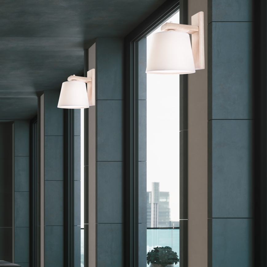 Nástenná lampa s bielym tienidlom je pripevnená na tmavej stene. V blízkosti je biela polica s váza s kvetmi. Kontext tvorí tmavá stena, zrkadlo a tmavé dvere.