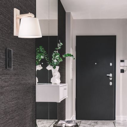 Biela textilná lampa sa pripája k drevenej konštrukcii. Lampa je pripevnená k svetlému drevenému podstavcu.