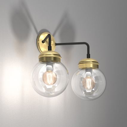 Dvojramenná nástenná lampa so sklenenými guľovými tienidlami a mosadznými prvkami je pripevnená na stene. Lampa má čierne kovové ramená. Svetlá sú zapnuté.