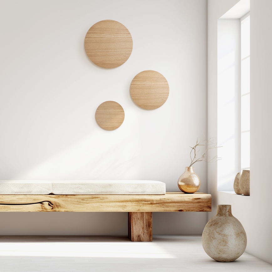 Drevená lavica s čalúneným sedákom stojí v svetlej miestnosti. Na stene sú tri okrúhle drevené dekoračné prvky. V blízkosti lavica sú dve vázy, jedna zlatá s konárikmi, druhá svetlohnedá.