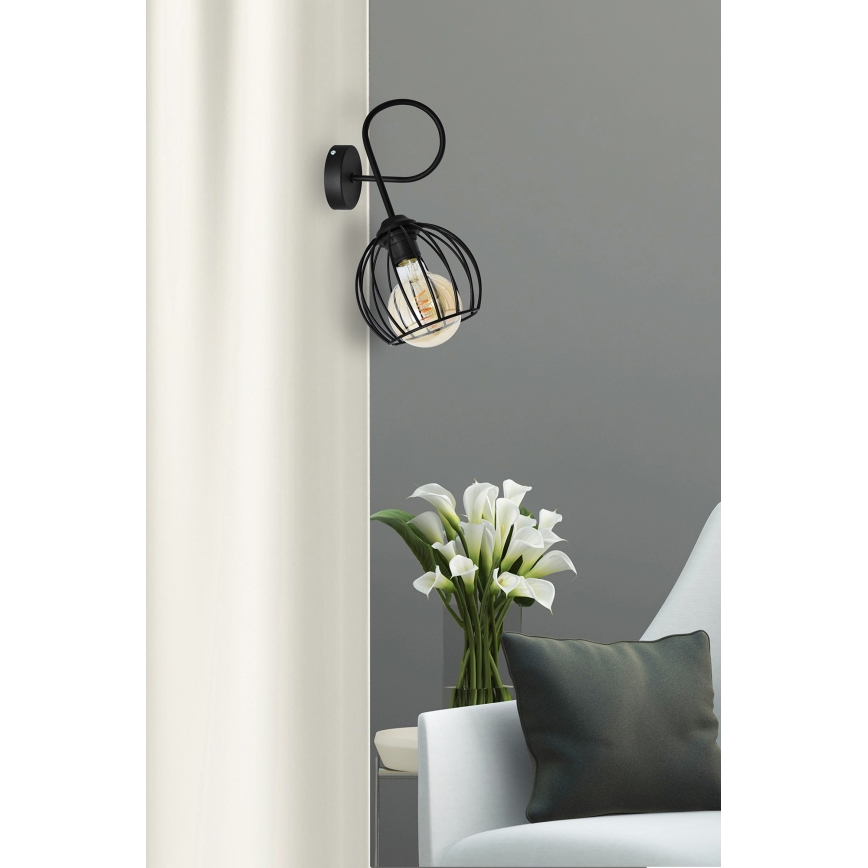 Čierna nástenná lampa s kovovým tienidlom v tvare gule svieti v interiéri. Lampa je pripevnená na stene vedľa svetlej záclony a pohovky s vázou bielych kvetov a tmavosivým vankúšom.