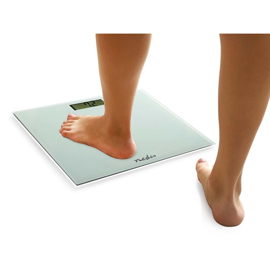 Noha stojí na digitálnych váhach s displejom ukazujúcim číslo 72. Váhy sú svetlo béžovej farby, s nápisom „nedis“. Druhá noha je mierne vzdialená od váh.