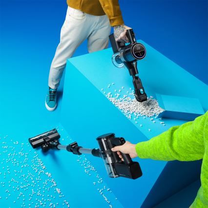 Dve bezdrôtové vysávače vysávajú drobné biele guľôčky na modrom podklade. Jedna osoba drží ručný vysávač, druhá používa tyčový.