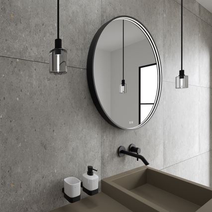 Okrúhle zrkadlo s podsvietením visí na stene v kúpeľni. Pri zrkadle je umývadlo a dávkovač mydla. Steny sú z betónu.