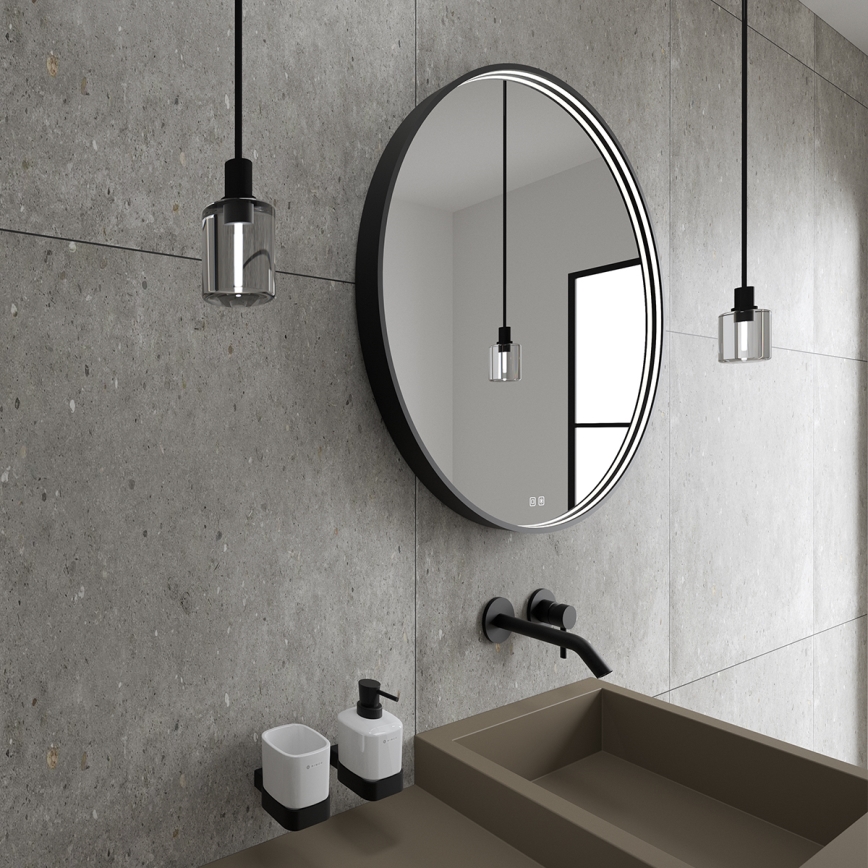 Okrúhle zrkadlo s podsvietením visí na stene v kúpeľni. Pri zrkadle je umývadlo a dávkovač mydla. Steny sú z betónu.