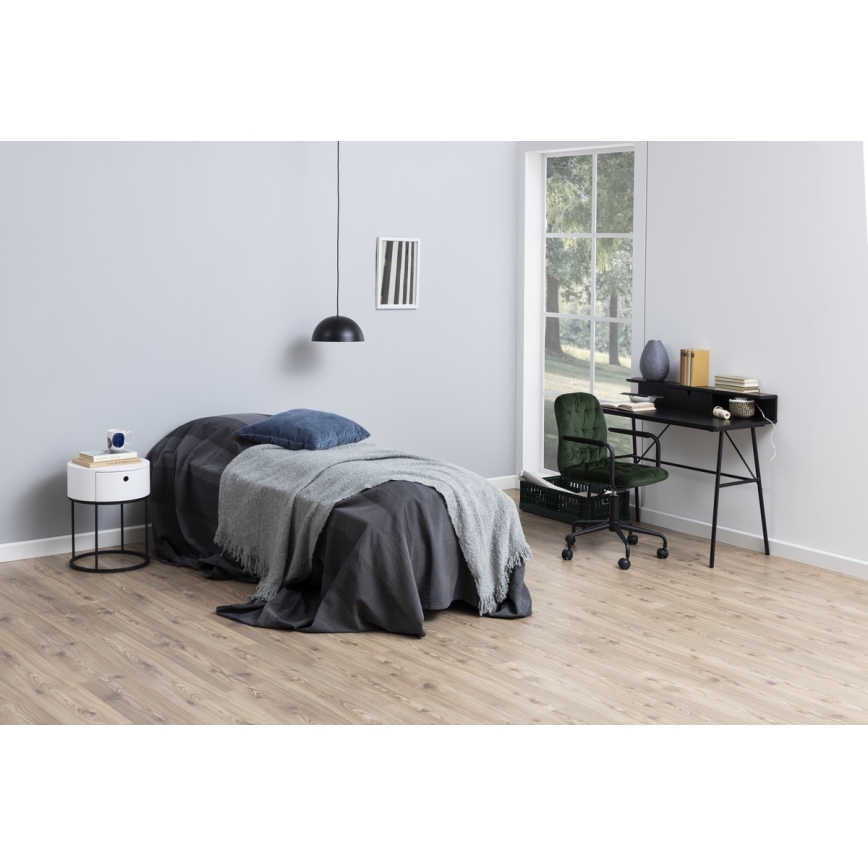 Posteľ s tmavosivou prikrývkou a vankúšom stojí v izbe s drevenou podlahou. Vedľa nej je nočný stolík. V pozadí je pracovný stôl s tmavozelenou kancelárskou stoličkou pri okne s výhľadom do záhrady.