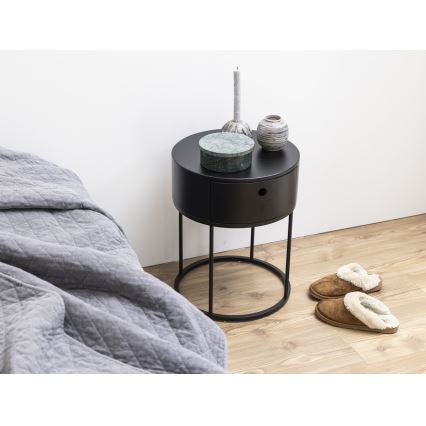 Nočný stolík s jedným šuplíkom stojí pri posteli. Na stolíku sú dekoratívne predmety: krabička, sviečka a vázy. Pri stolíku sú papuče.