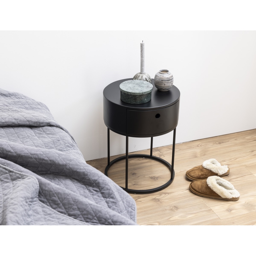 Nočný stolík s jedným šuplíkom stojí pri posteli. Na stolíku sú dekoratívne predmety: krabička, sviečka a vázy. Pri stolíku sú papuče.