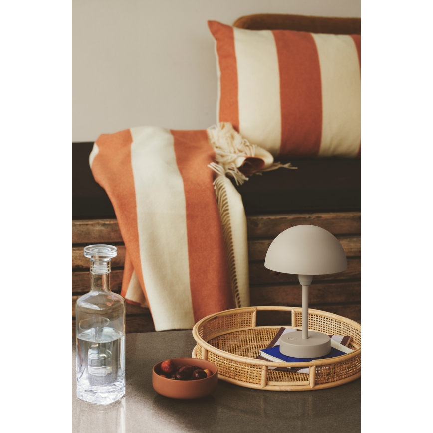 Stolová lampa s polguľovitým tienidlom stojí na ratanovom podnose. Na podnose sú aj knihy. V pozadí je drevený nábytok s oranžovo-bielou dekou a vankúšom.