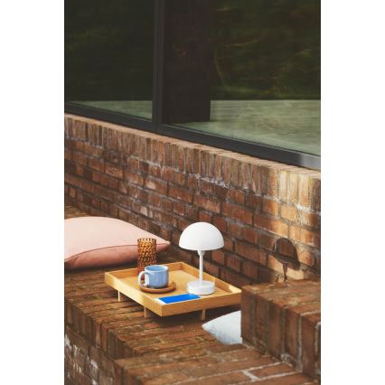 Biela stolová lampa s kupolou stojí na drevenom podnose. Na podnose je šálka, podšálka a kniha. Kontext tvorí tehlová stena a sedacia časť.