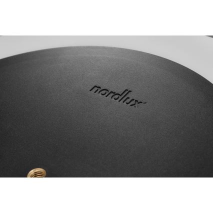Čierna okrúhla platňa s vygravírovaným logom „nordlux®“ a zlatým skrutkovým otvorom je zobrazená na svetlom pozadí.