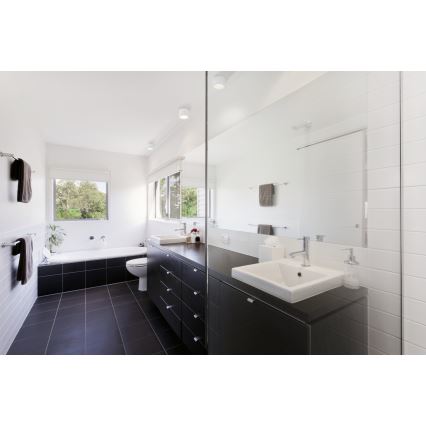 Moderná kúpeľňa s umývadlom a vaňou. V priestore dominuje tmavá nábytková zostava s umývadlom a vstavanými skrinkami. Kúpeľňa je obložená bielymi a tmavými dlaždicami. Okno poskytuje výhľad na zeleň.