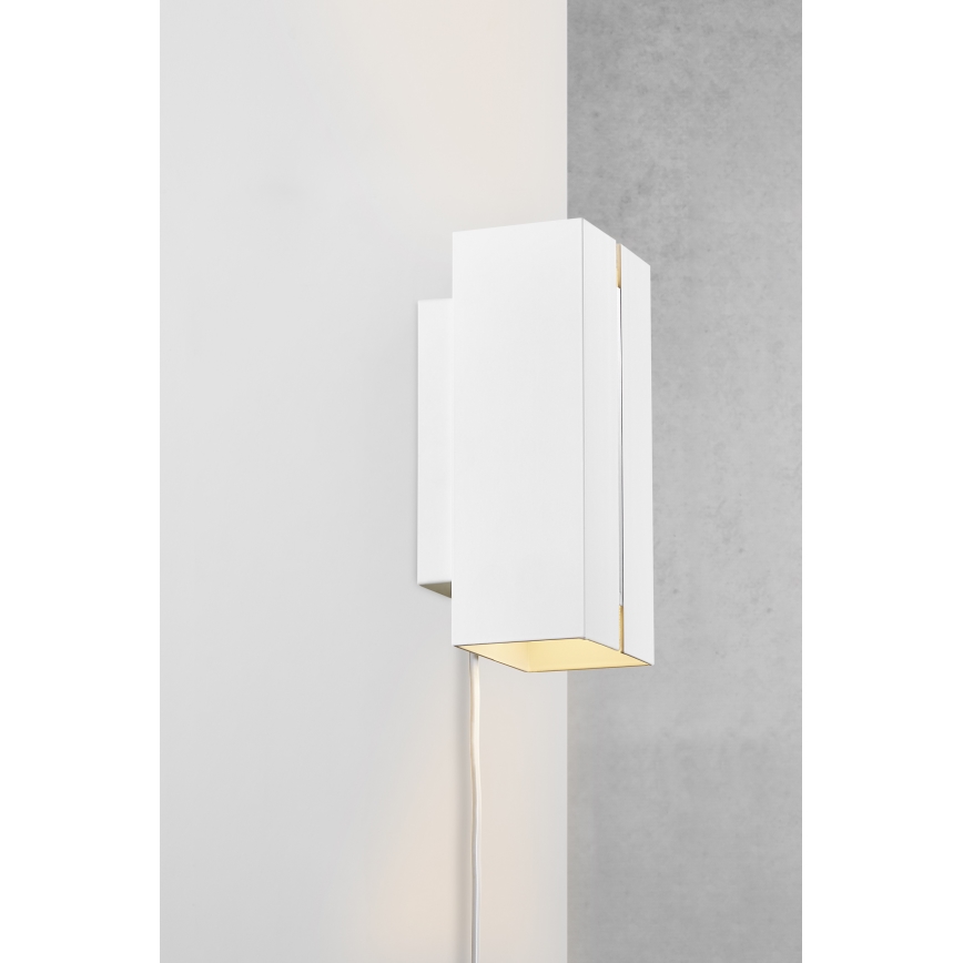 Biela nástenná lampa svieti. Je pripevnená na stene, vedľa sivej steny. Svetlo vychádza z jej spodnej časti.