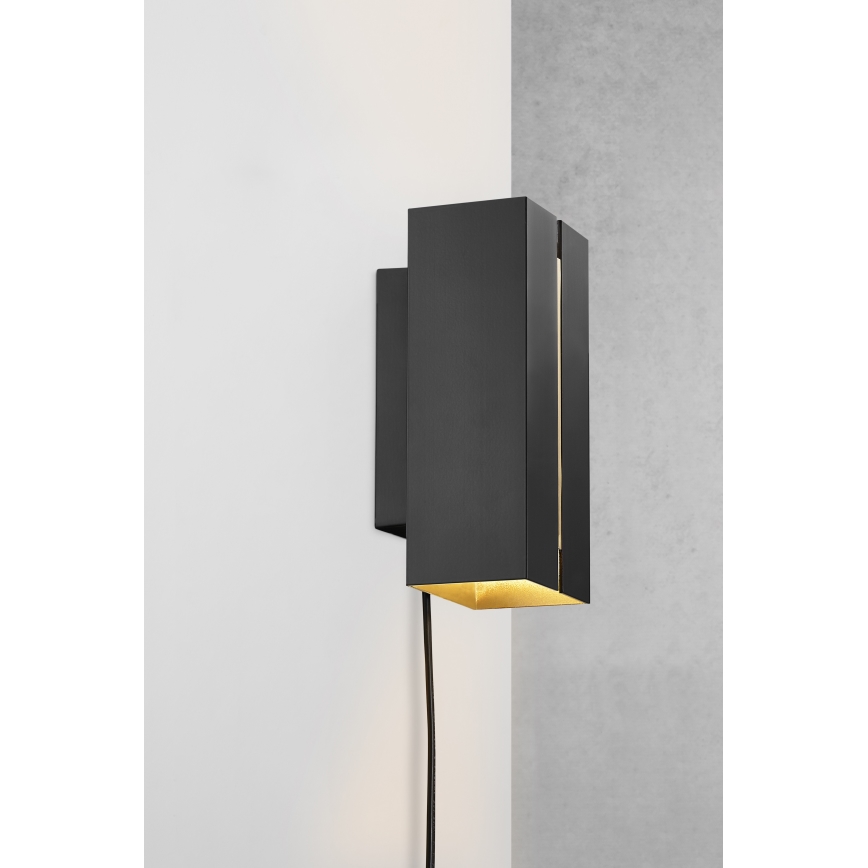 Čierna nástenná lampa s hranatým dizajnom svieti. Je pripevnená na svetlej a tmavej stene. Vidieť je aj čierny napájací kábel.