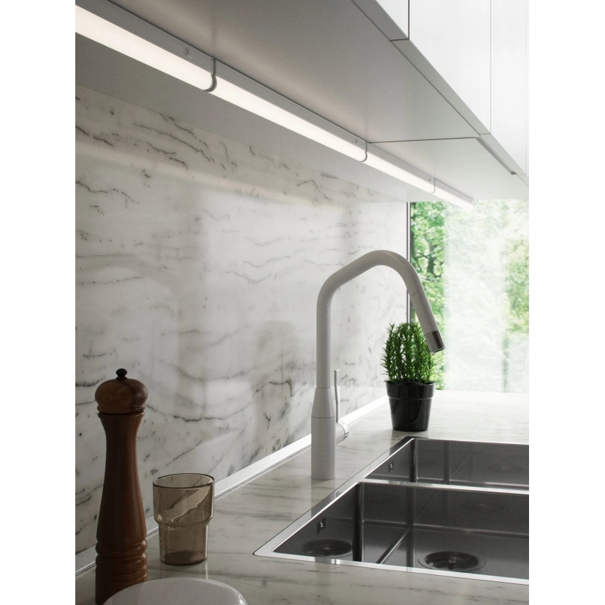 LED pásik osvetľuje kuchynskú linku s mramorovou stenou. Pásik je pripevnený pod hornými skrinkami nad drezom a pracovnou doskou. V popredí je vidieť drez, batériu a kuchynské doplnky.