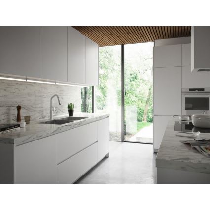 Dvojzákladňový drez s batériou je umiestnený na kuchynskej linke z bieleho materiálu s mramorovým povrchom. Linka je súčasťou modernej kuchyne s veľkým oknom s výhľadom do záhrady.