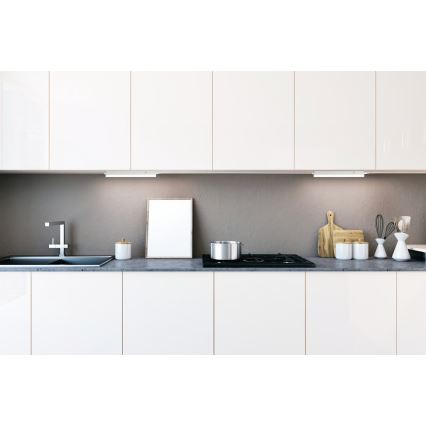 Kuchynská linka s bielymi skrinkami a sivou pracovnou doskou. Na doske je umiestnený hrniec, nádoba, rám obrazu a kuchynské náčinie. Pod skrinkami je osvetlenie.